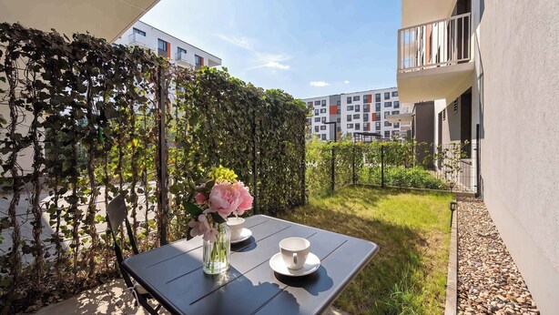 A table with a vase of flowers and two cups, a hedge, and a building in the background. - Cozy Studio with a Garden | A Place to Work (Wrocław)