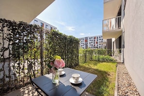 A table with a vase of flowers and two cups, a hedge, and a building in the background. - Cozy Studio with a Garden | A Place to Work (Wrocław)