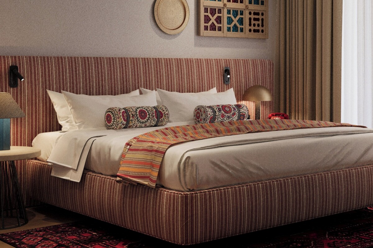 Habitación de hotel con una cama grande, cabecera a rayas, almohadas estampadas, una mesita de noche con lámpara y un elemento decorativo en la pared.