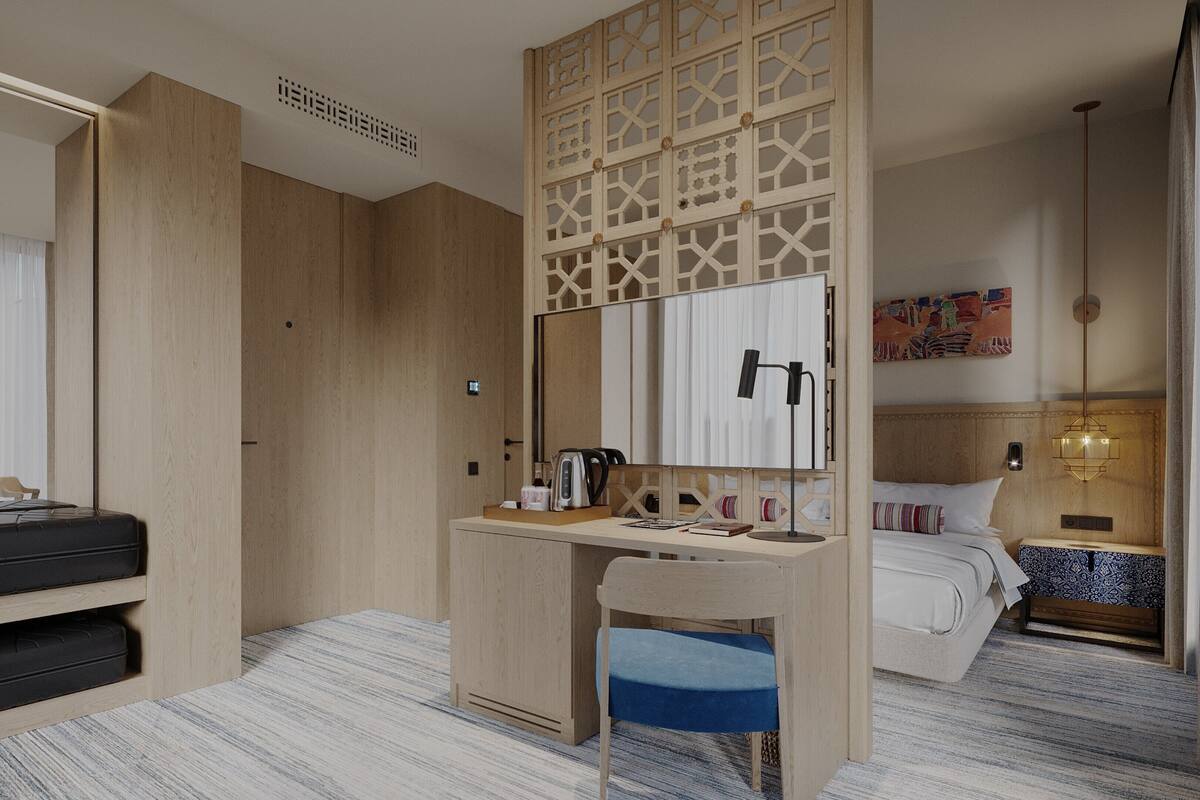 A hotel room with a bed, a desk with a mirror, a chair, a suitcase, and a bedside table with a lamp.