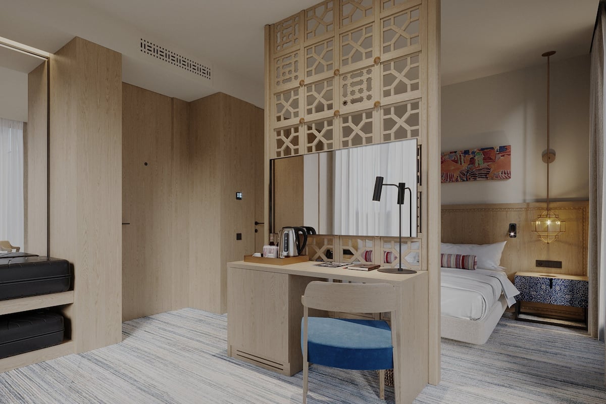 A hotel room with a bed, a desk with a mirror, a chair, a suitcase, and a bedside table with a lamp.