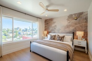 A bedroom with a bed, nightstands, a ceiling fan, a large window with a city view, and a textured wall.