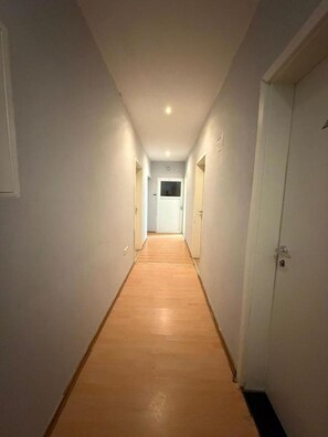 Un corridoio stretto con pavimento in legno e pareti bianche, che conduce a una porta in fondo.