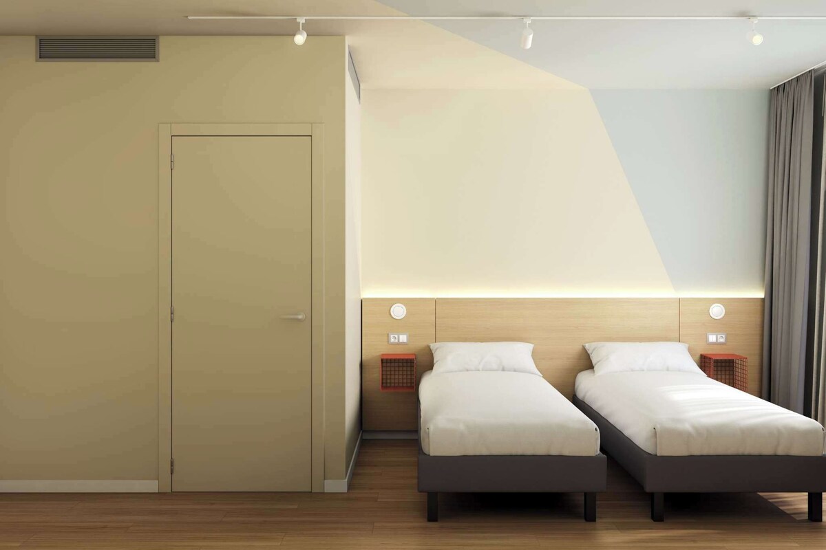 A modern hotel room with two single beds, a wardrobe, and a large window with curtains.