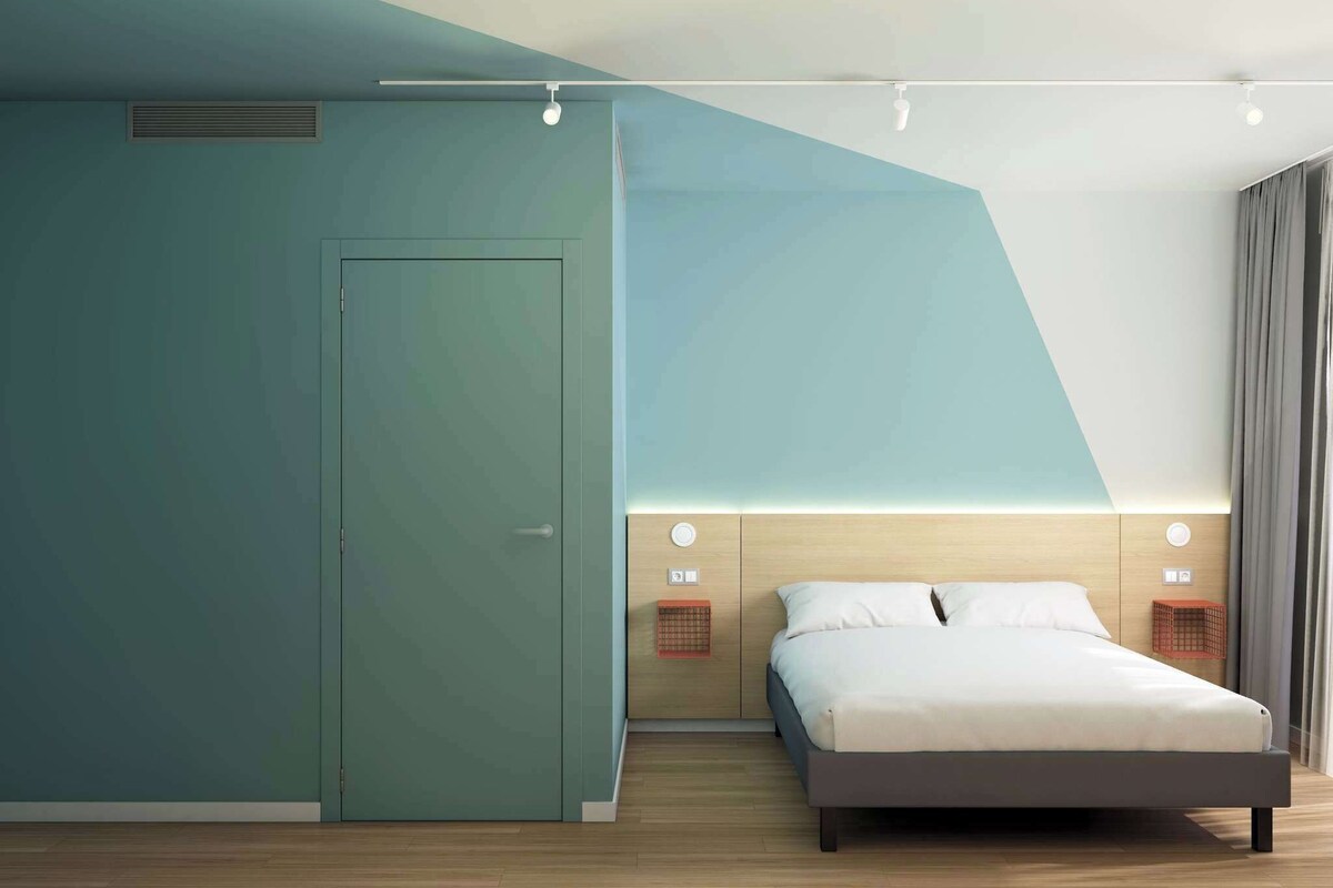 A modern hotel room with a bed, a wardrobe, a headboard with a light fixture, and a wall-mounted heater.
