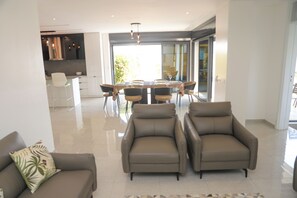 Un salon moderne avec un canapé, deux fauteuils, une table à manger avec des chaises, et une cuisine en arrière-plan.