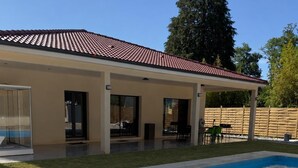 Una piscina con solárium de baldosas, un porche cubierto y una valla de madera al fondo.
