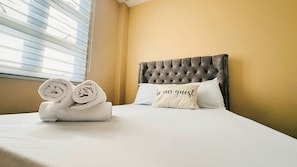 Una cama bien hecha con toallas enrolladas y una almohada con el texto 'be our guest', un cabecero capitonado y una ventana con persianas.