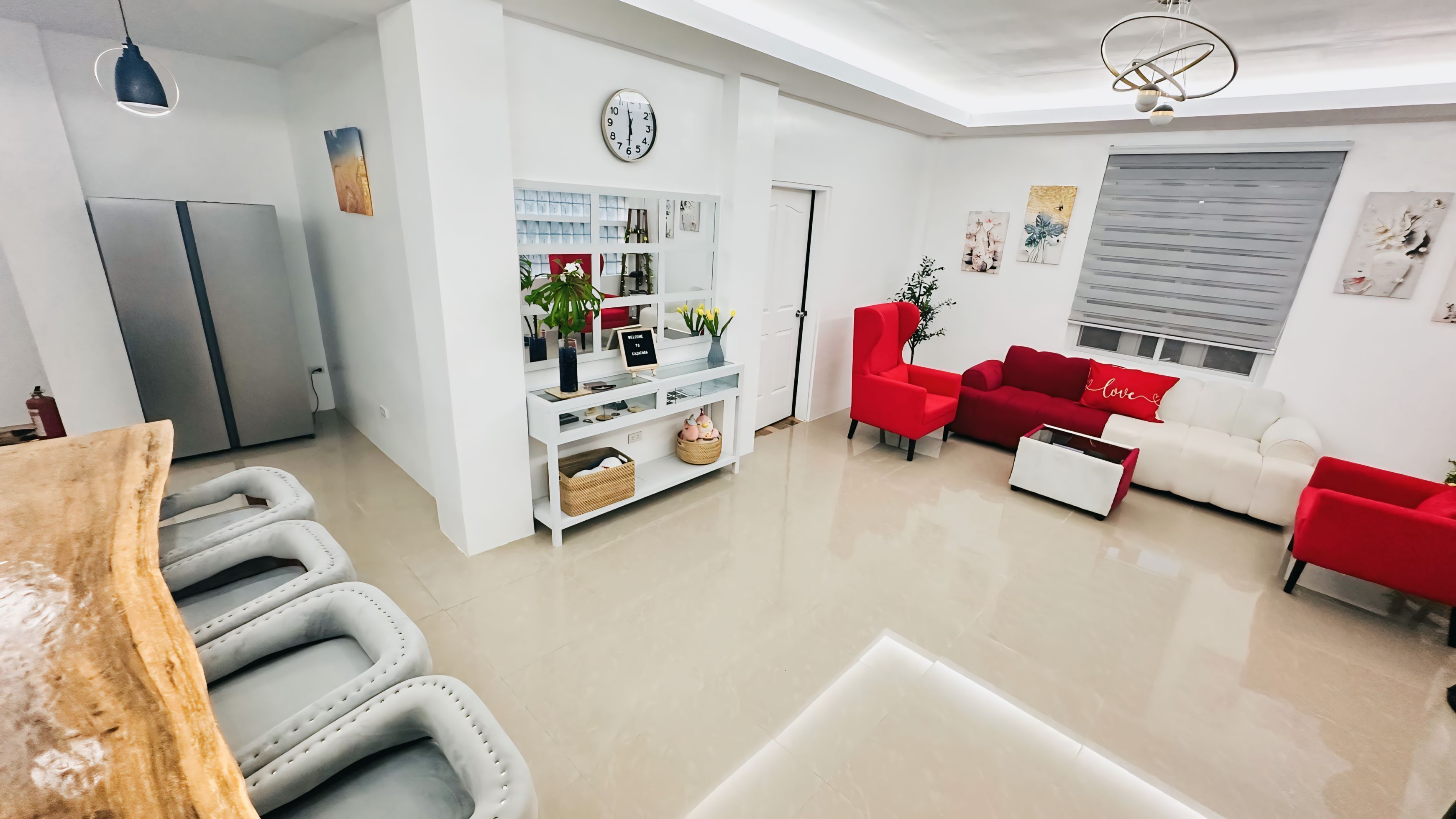 A modern living room with a red sofa, a white sofa, and a red armchair. There is a kitchen area with a refrigerator and a white cabinet. A clock is visible on the wall, and a plant is on the floor.