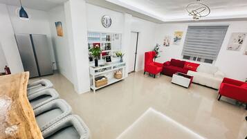 A modern living room with a red sofa, a white sofa, and a red armchair. There is a kitchen area with a refrigerator and a white cabinet. A clock is visible on the wall, and a plant is on the floor.