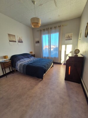 Ein Schlafzimmer mit Bett, Nachttisch, Kommode, einem Fenster mit Vorhängen und Wandbildern.