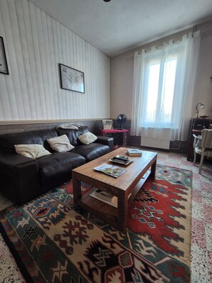 A living room with a leather sofa, a wooden coffee table, a patterned rug, a framed picture on the wall, and a window with curtains. - Fourasine family home 300m from the beach (Fouras)
