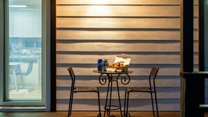 Una mesa con un cubo de pollo frito, una lata de cerveza y un recipiente pequeño de salsa, frente a una pared de madera con una ventana al fondo.