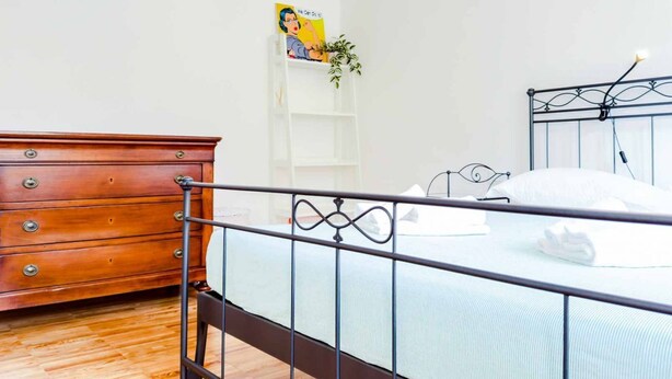 Une chambre avec une commode en bois, un cadre de lit en métal, un mur blanc et une petite étagère sur laquelle est posée une plante.