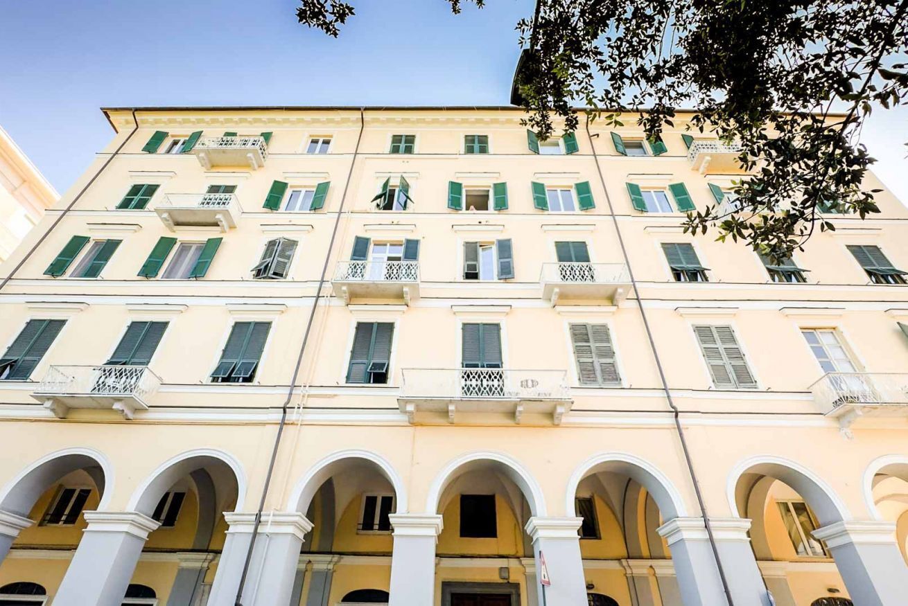 Un edificio de varios pisos con fachada amarilla, contraventanas verdes y columnas blancas que sostienen una entrada en arco.