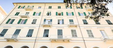 Un immeuble à plusieurs étages, doté d’une façade jaune, de volets verts et de colonnes blanches soutenant un porche en arc de cercle.