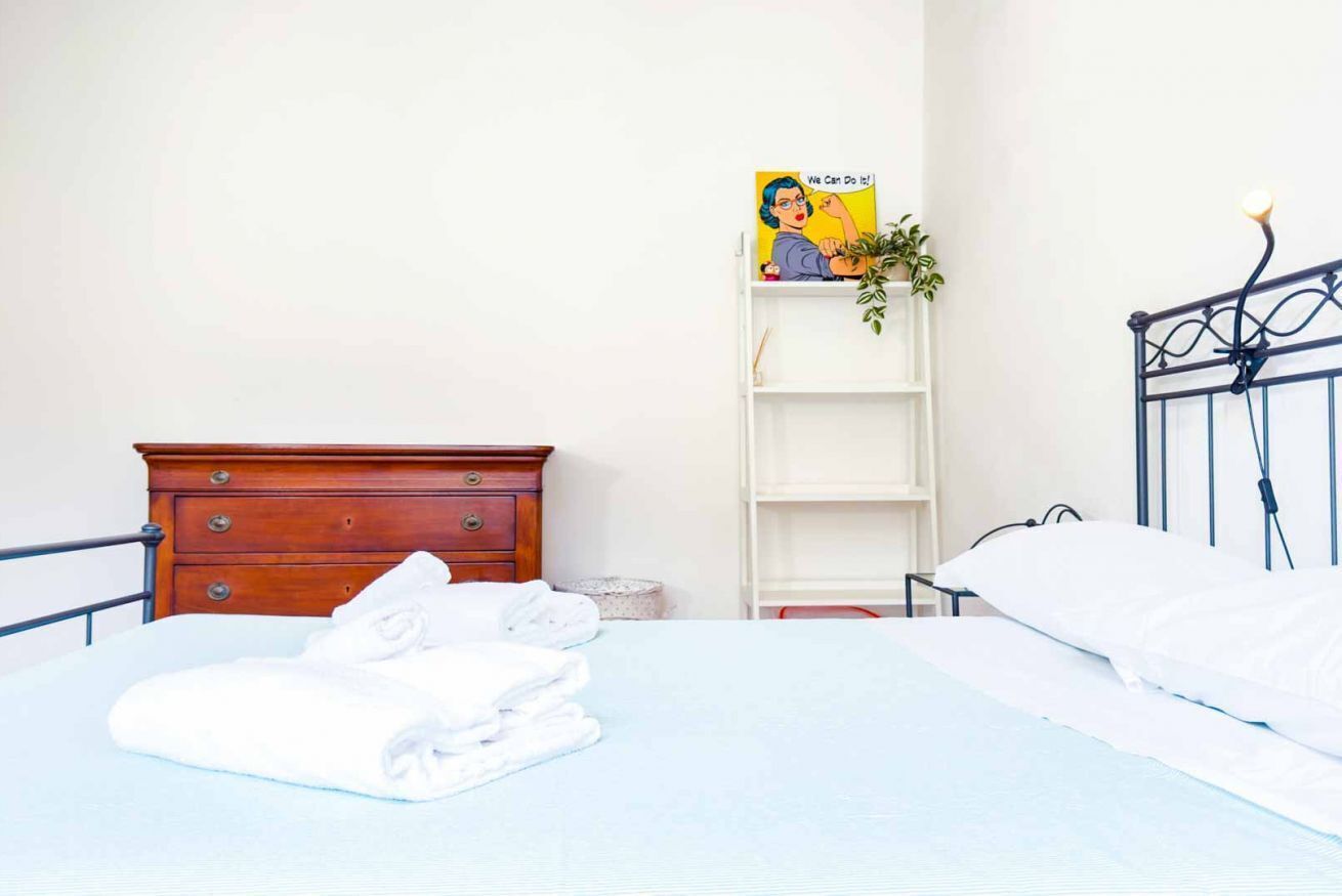Un letto ben rifatto con lenzuola bianche, un comò in legno, una mensola bianca con una pianta e un poster appeso al muro.
