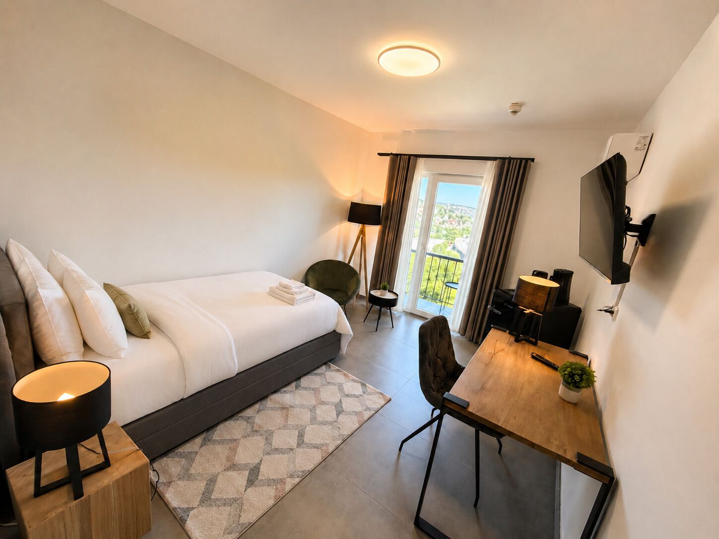 Una habitación de hotel moderna con cama, mesita de noche, lámpara, escritorio, silla, televisor y vistas al exterior.
