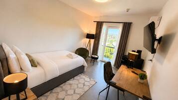 Una habitación de hotel moderna con cama, mesita de noche, lámpara, escritorio, silla, televisor y vistas al exterior.