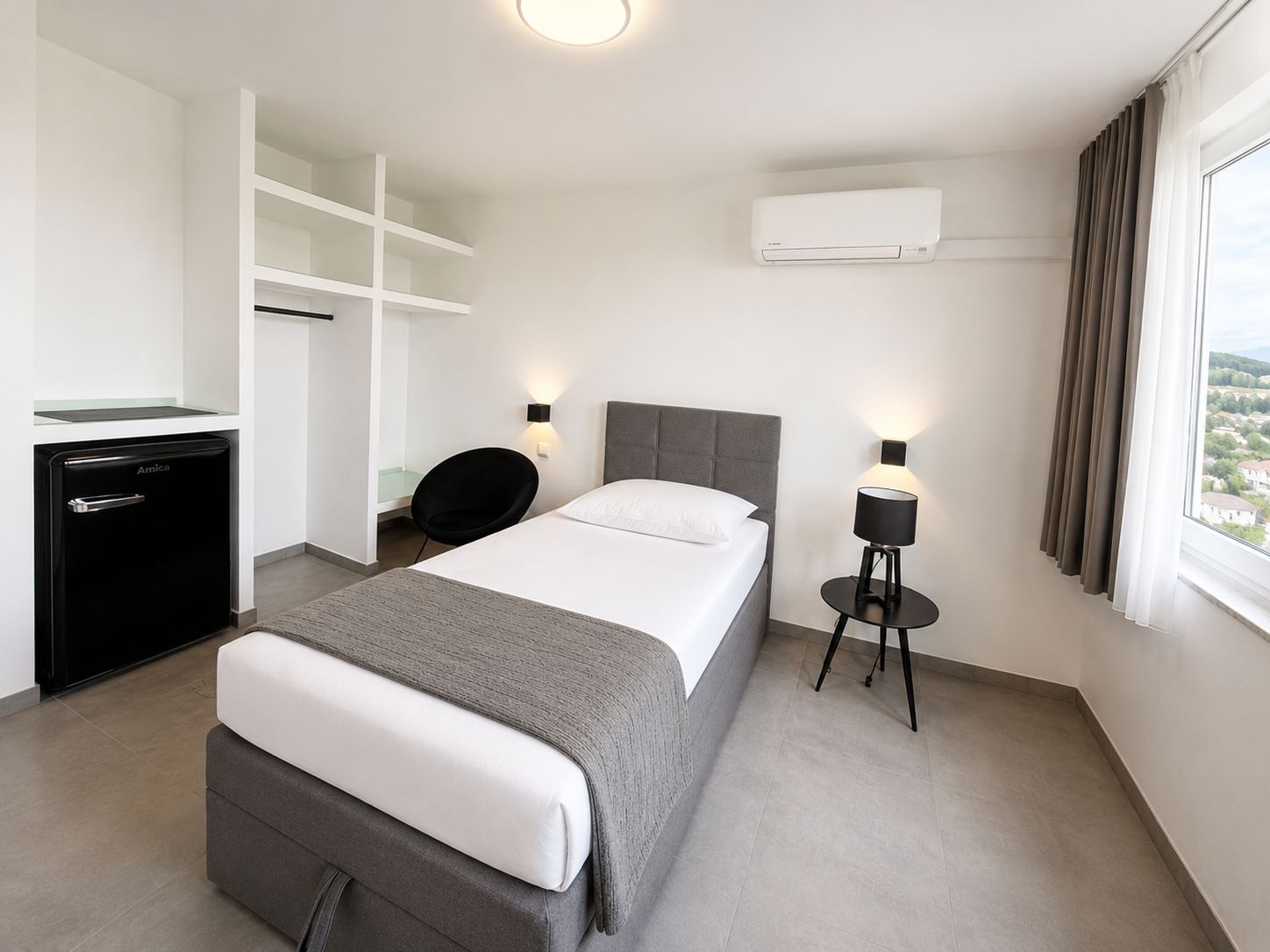 A hotel room with a bed, a desk, a chair, a lamp, and an air conditioning unit.