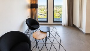 Una habitación de hotel moderna con un ventanal grande, dos sillas negras, una mesita de madera y una alfombra con diseño geométrico.