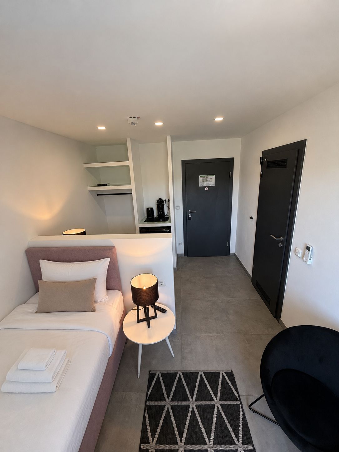 Una habitación de hotel moderna con una cama, una mesita con una lámpara, una silla negra, una puerta oscura y una pared blanca con un estante.