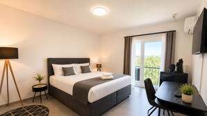 Una habitación de hotel moderna con una cama, un escritorio, una silla, una mesita, una lámpara, una planta en maceta y vistas al exterior.