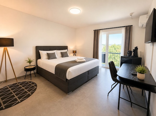 A modern hotel room with a bed, a desk, a chair, a small table, a lamp, a potted plant, and a view of the outdoors. - Elion Hotel Siegen (Siegen)