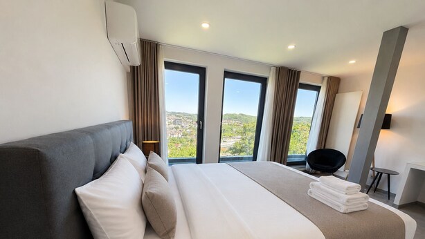 Habitación de hotel moderna con una cama grande, una silla y vistas a un paisaje urbano a través de las ventanas.