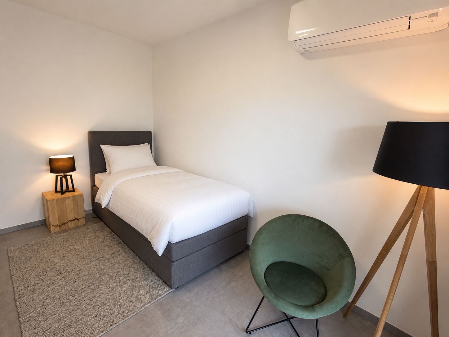 A bedroom with a bed, a bedside lamp, a green chair, and an air conditioning unit.