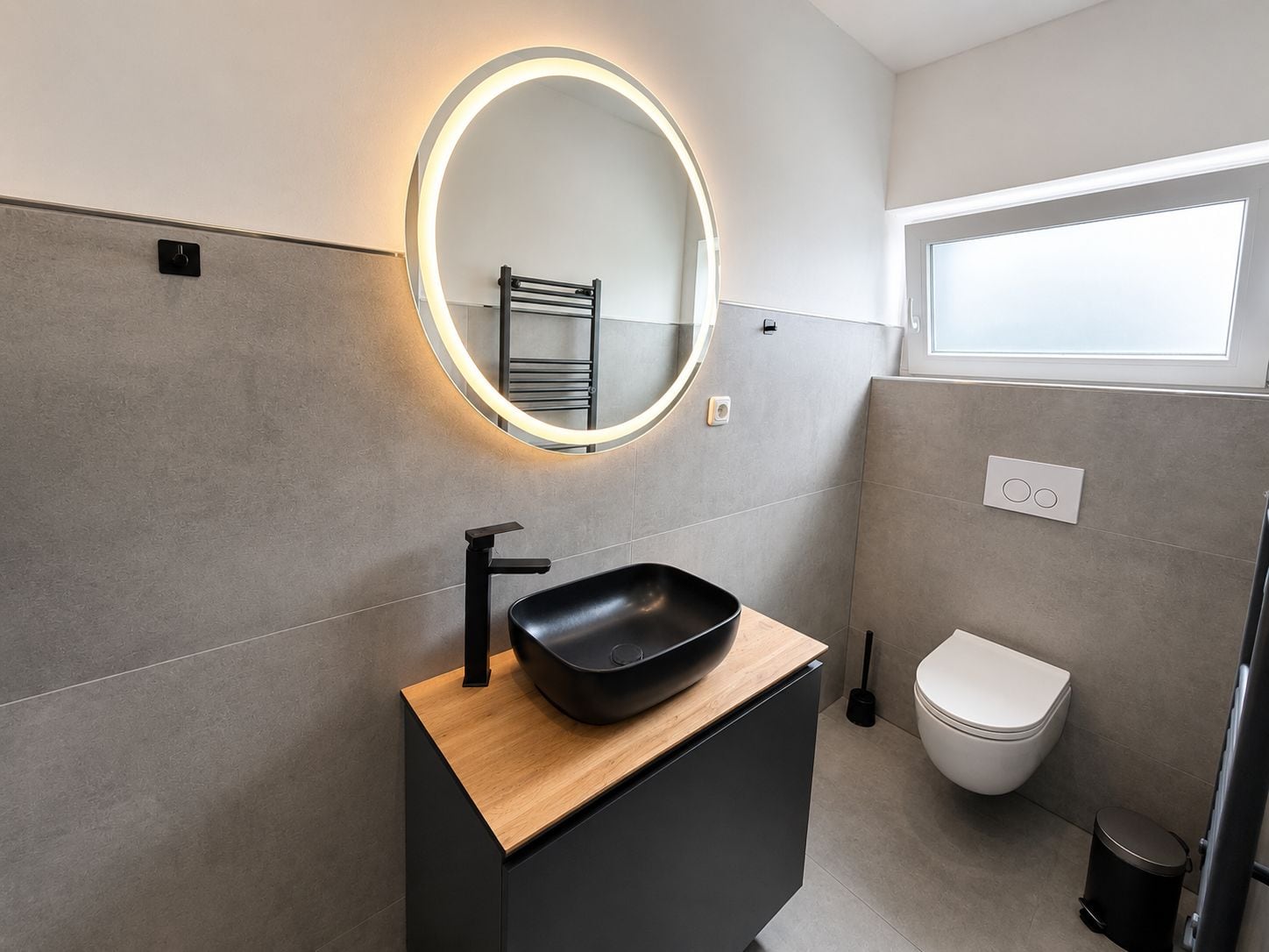 Un baño moderno con un lavamanos negro, un espejo redondo con iluminación, un inodoro y un ventanal.
