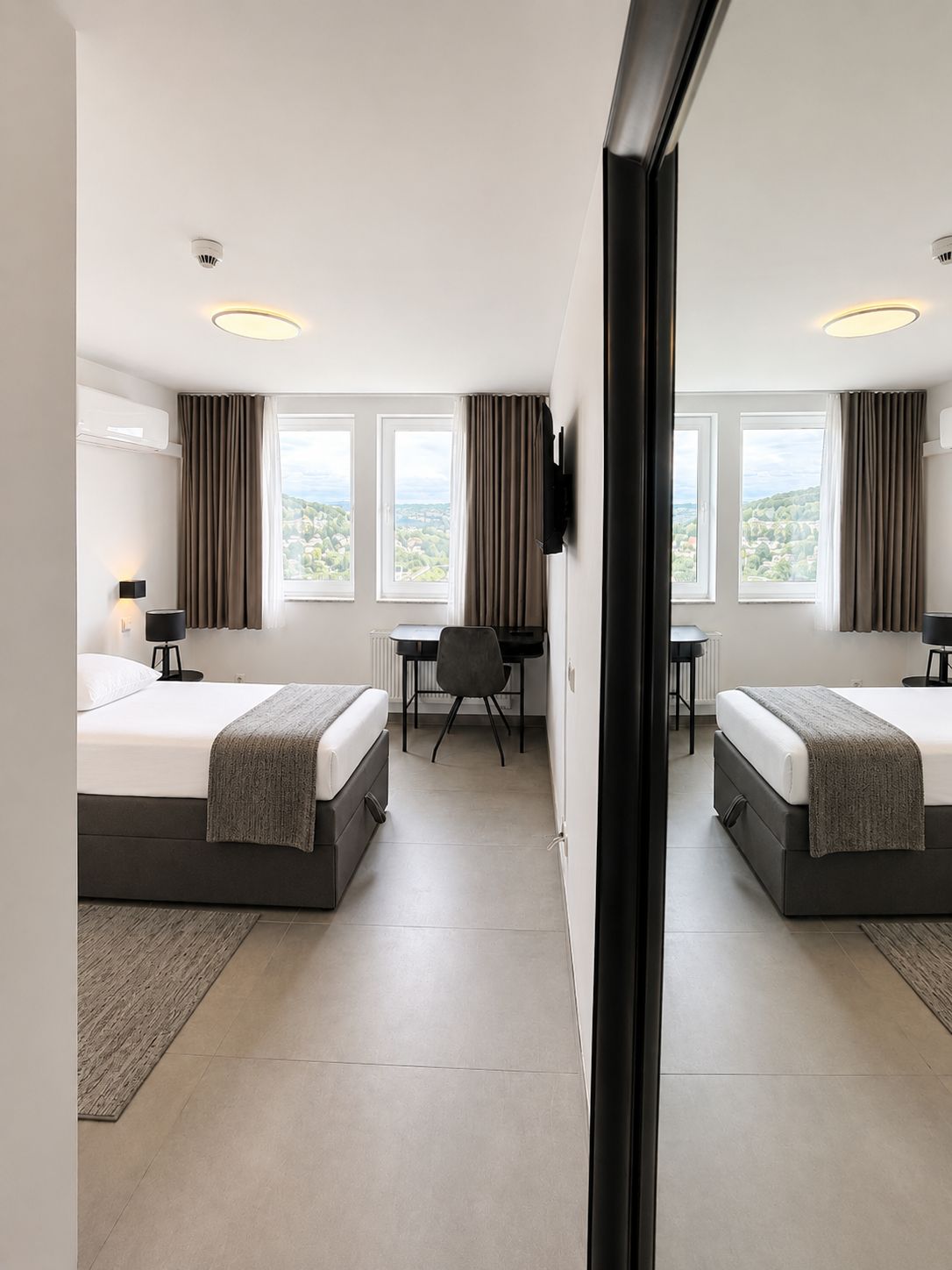 Habitación de hotel con dos camas, un escritorio y una silla. Hay ventanas con cortinas, un televisor de pantalla plana montado en la pared y un equipo de aire acondicionado.