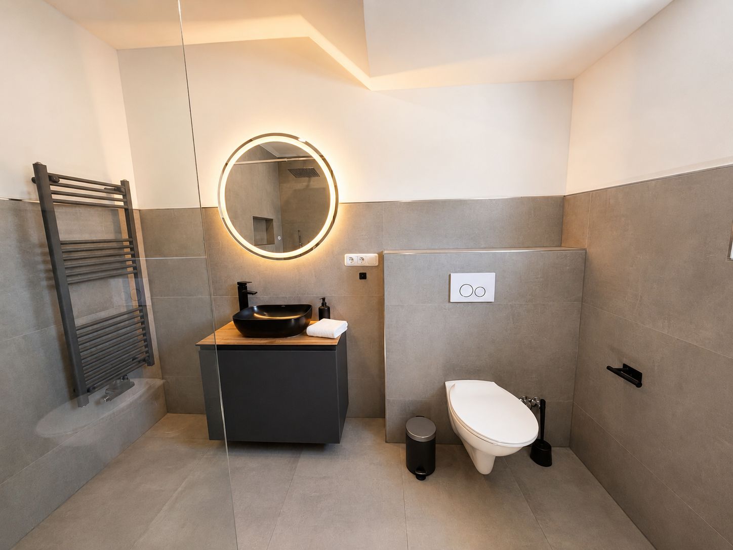 Ein modernes Badezimmer mit einem schwarzen Waschbecken, einem runden Spiegel mit Beleuchtung, einer Toilette und einem Handtuchhalter.