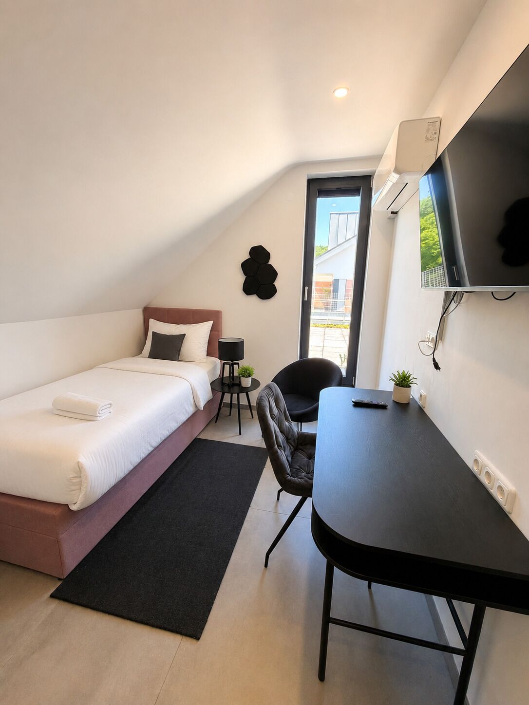 A modern bedroom with a bed, a desk, a chair, a small table, a potted plant, and a TV mounted on the wall.