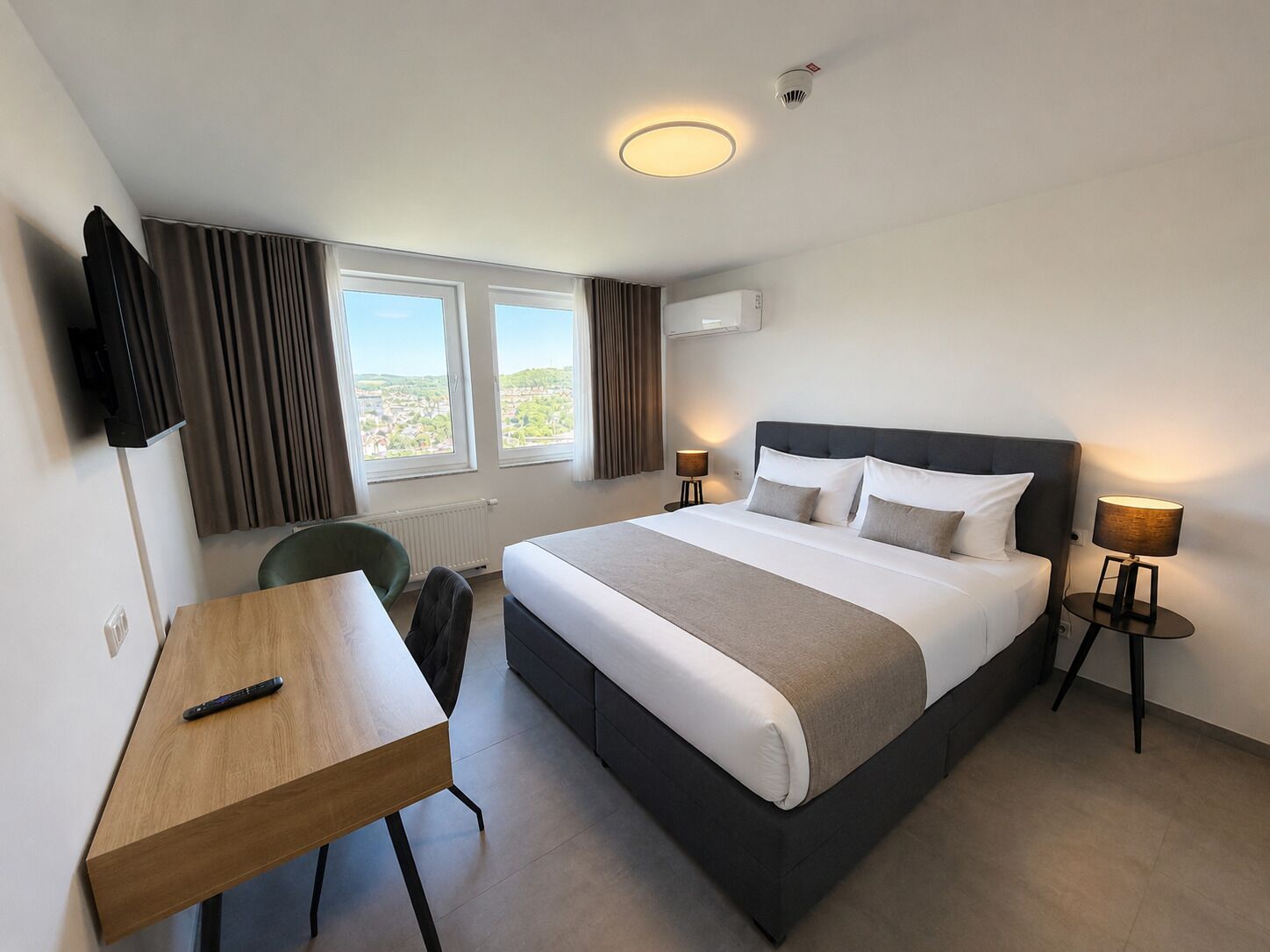 Habitación de hotel con cama, mesitas de noche, escritorio, silla, televisor y ventana con cortinas.