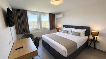 Habitación de hotel con cama, mesitas de noche, escritorio, silla, televisor y ventana con cortinas.