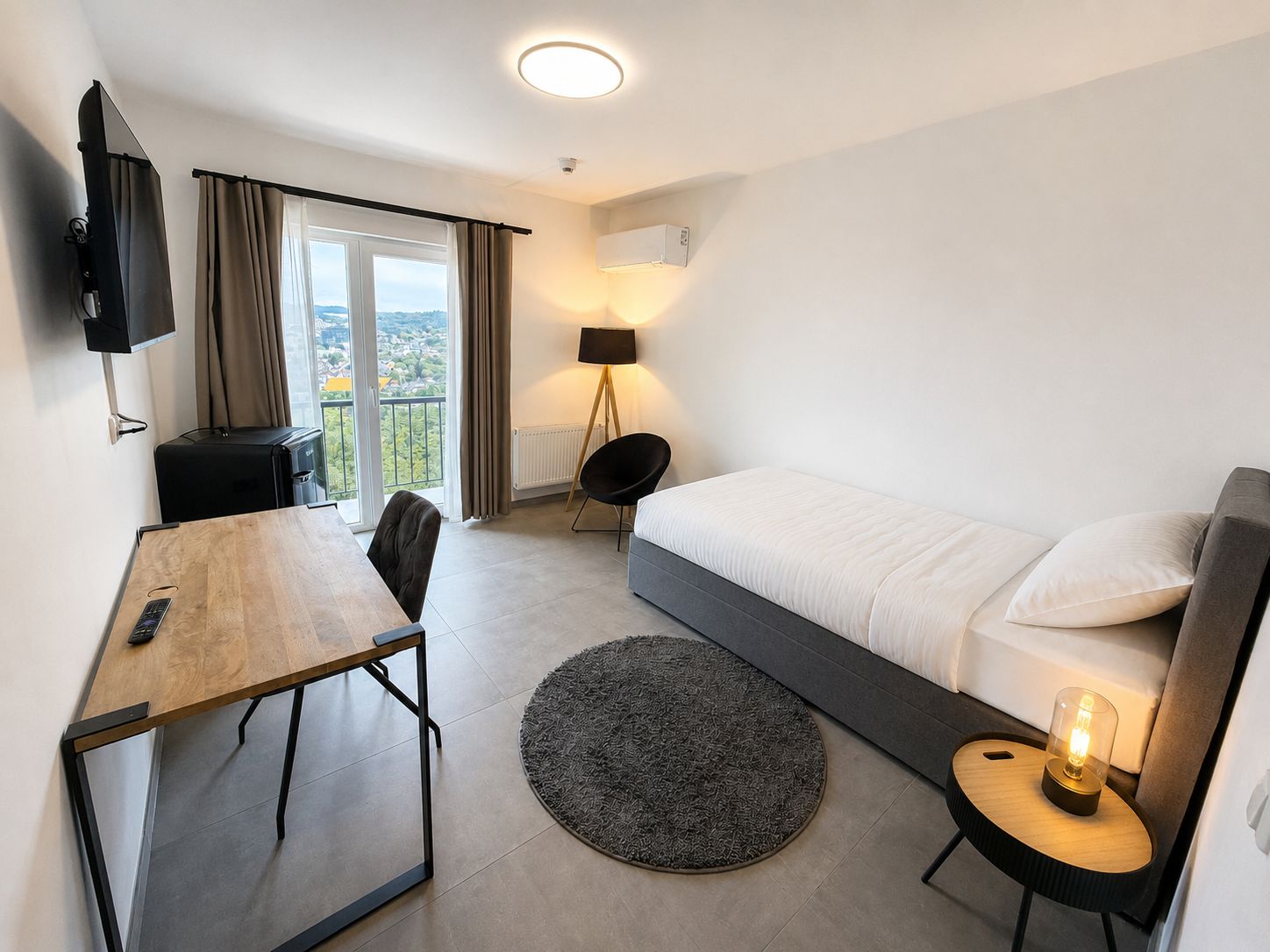Ett hotellrum med en säng, ett skrivbord med en stol, en golvlampa, en rund matta och en tv som är monterad på väggen.