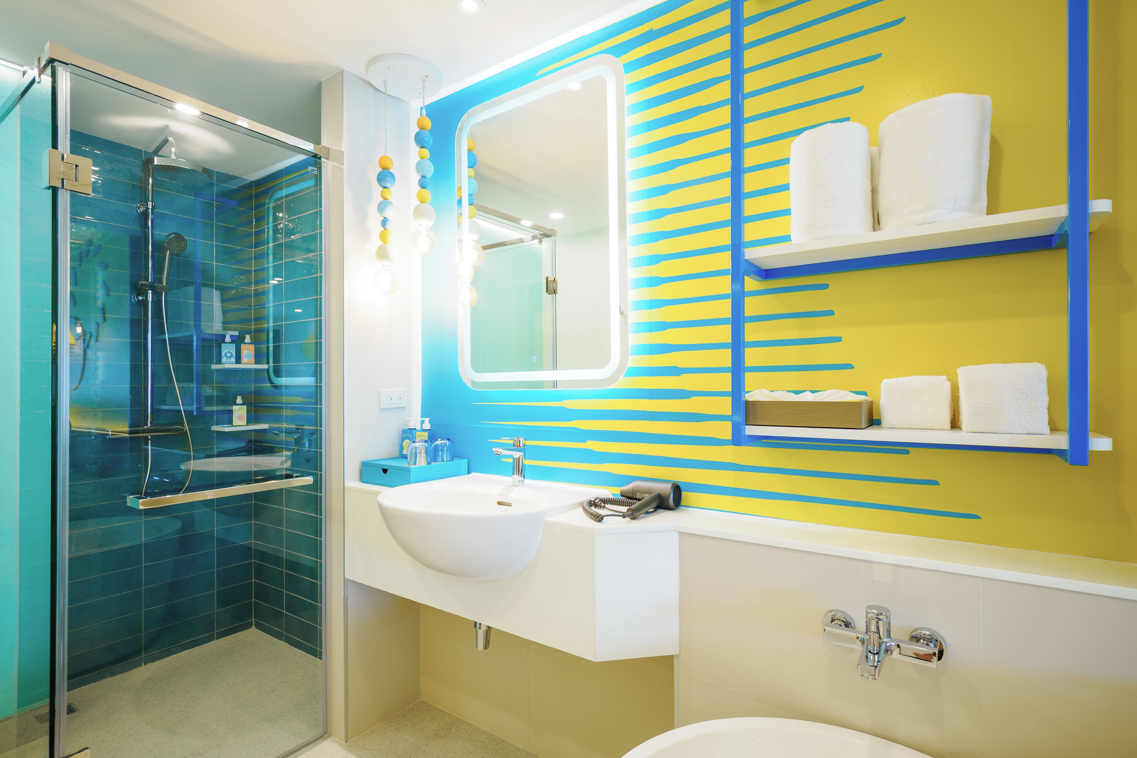 A modern bathroom with a glass shower enclosure, a white sink, a large mirror, and a yellow and blue striped wall.