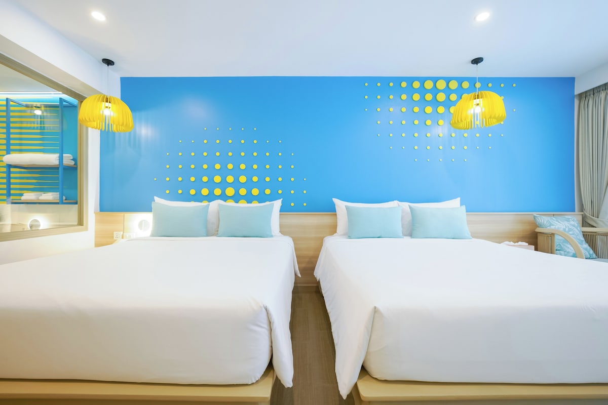 Een hotelkamer met twee bedden, een blauwe accentmuur en een gele hanglamp.