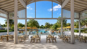 Un área de piscina techada al aire libre con mesas y sillas, una vista despejada de la piscina y palmeras al fondo.