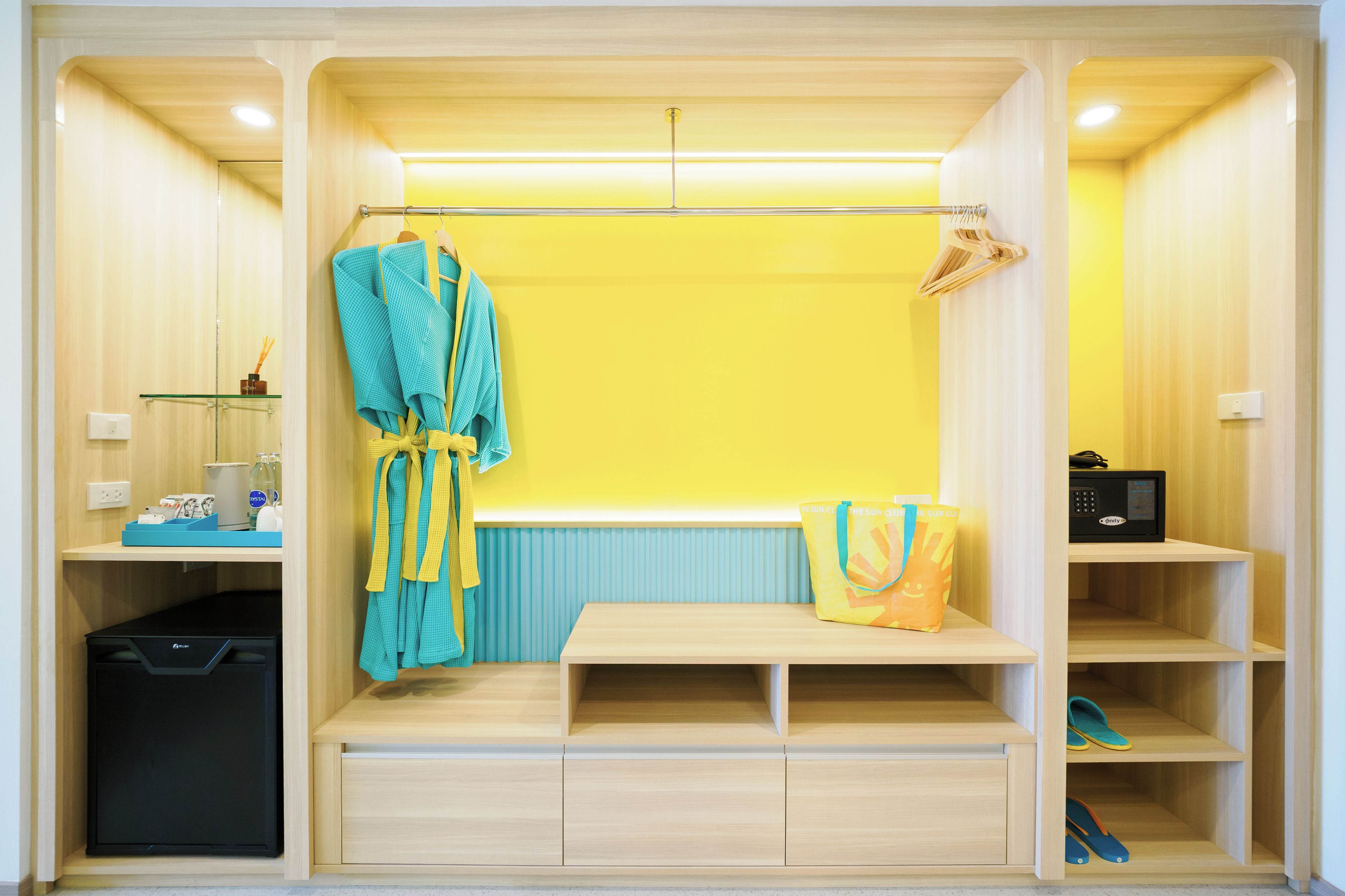 A compact room with a bench, a robe, a bag, and a shelf with slippers.