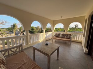 A covered balcony with a table and seating, white railings, and a view of greenery.
