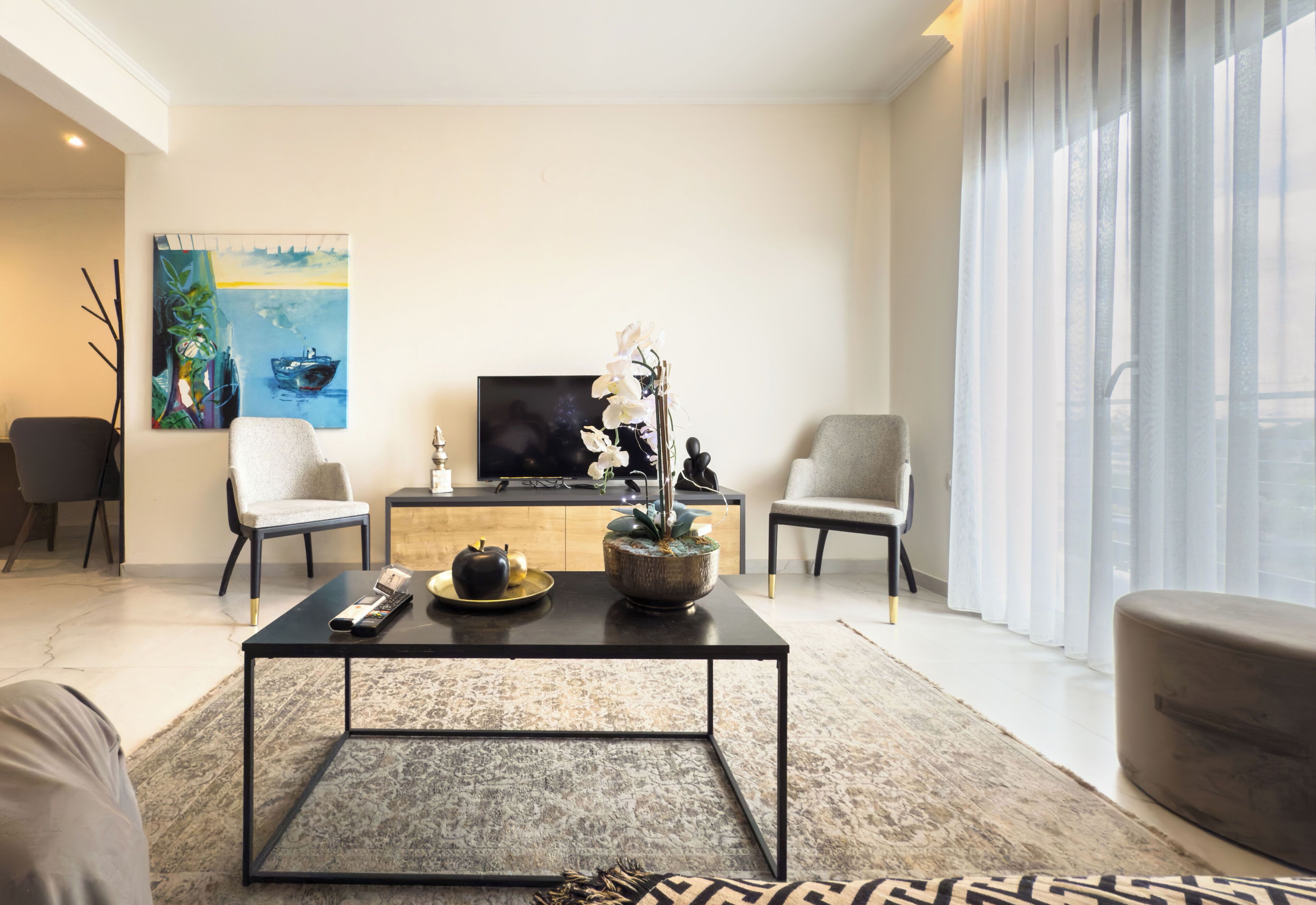 A modern living room with a flat-screen TV, a coffee table, two armchairs, a sofa, and a large window with sheer curtains.
