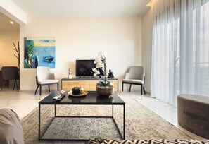 A modern living room with a flat-screen TV, a coffee table, two armchairs, a sofa, and a large window with sheer curtains. - Alexandra Boutique Residence Downtown  (Alexandroupoli)