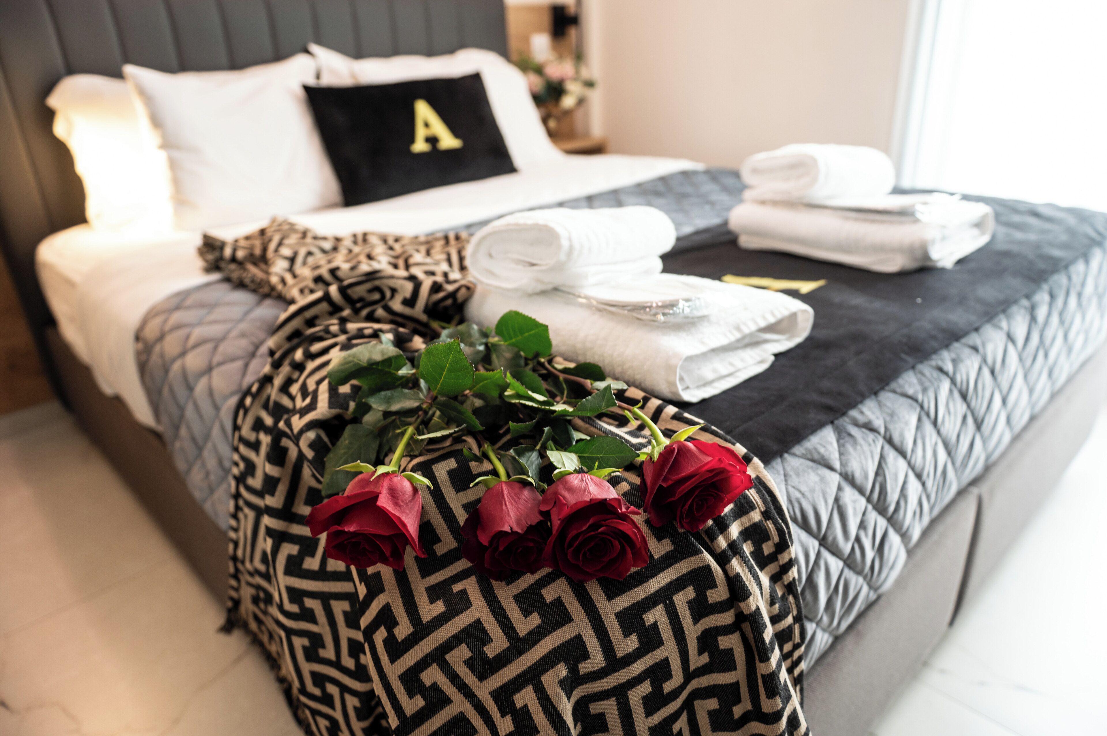 Ein ordentlich bezogenes Bett mit einem Kopfkissen mit dem Buchstaben 'A', einer gemusterten Tagesdecke, gefalteten Handtüchern und einem Rosenstrauß in Rot.