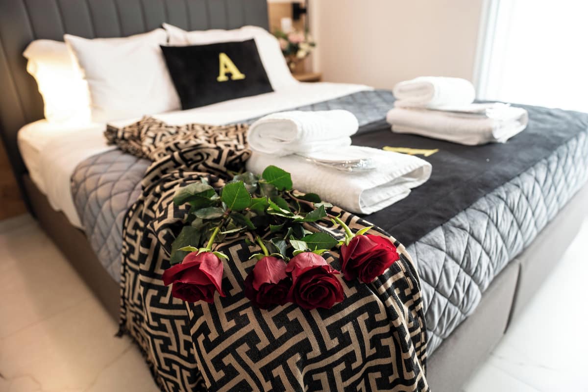 A neatly made bed with a pillow marked with an 'A', a patterned throw, folded towels, and a bouquet of red roses.