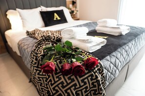 Ein ordentlich bezogenes Bett mit einem Kopfkissen mit dem Buchstaben 'A', einer gemusterten Tagesdecke, gefalteten Handtüchern und einem Rosenstrauß in Rot.