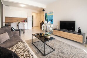 Ein modernes Wohnzimmer mit einer Couch, einem Couchtisch und einem Fernseher. Im Hintergrund ist eine Küche zu sehen.