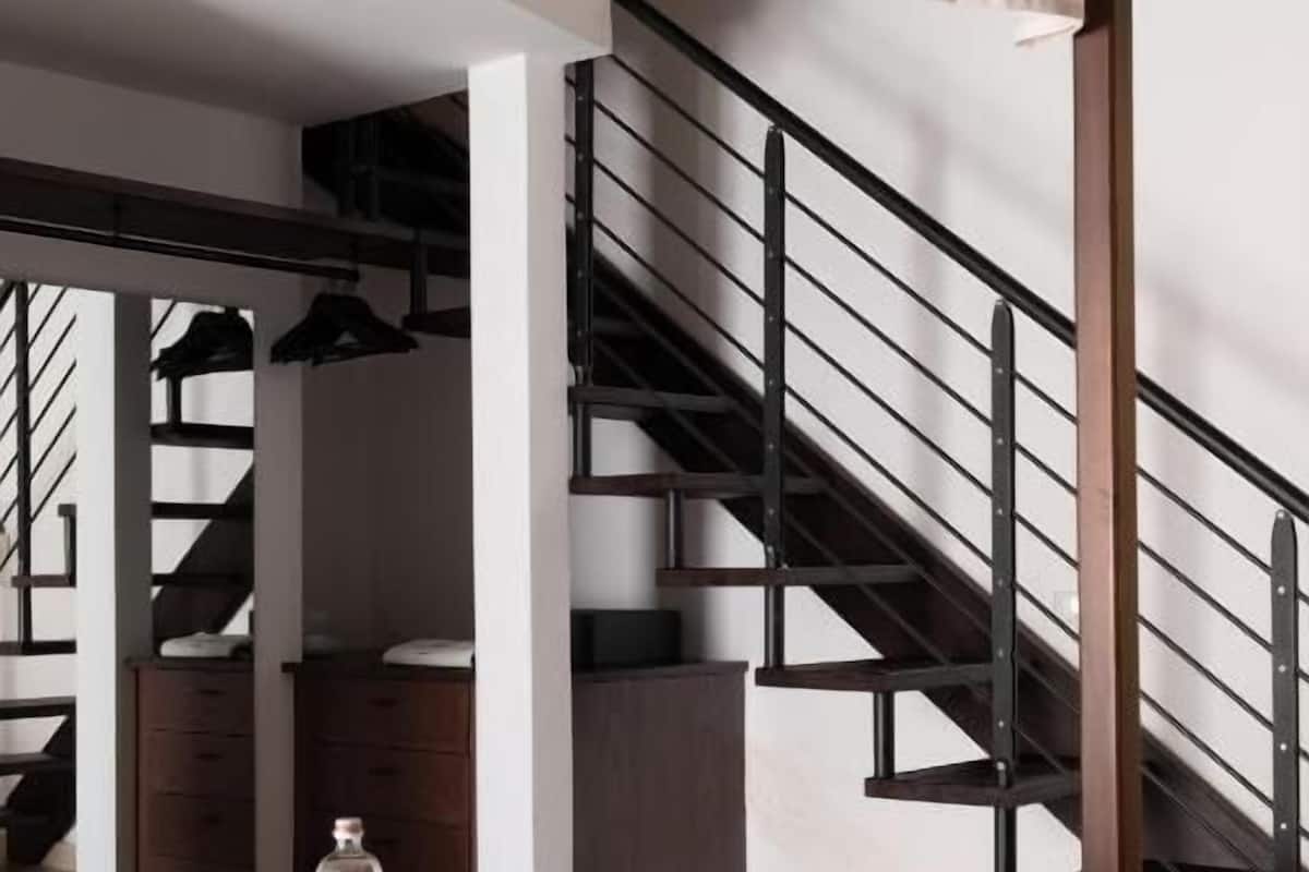 A bedroom with a bed, a nightstand, a glass of water, a bottle, a chair, a table, and a staircase.