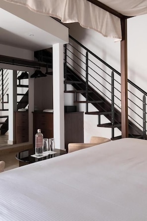 Quarto com cama, mesa de cabeceira, copo de água, garrafa, cadeira, mesa e escada.